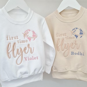 May include: Two white and beige sweatshirts with the text "first time flyer" embroidered on them. The sweatshirts have a globe and airplane design and the names "Violet" and "Bodhi" are embroidered below the text.