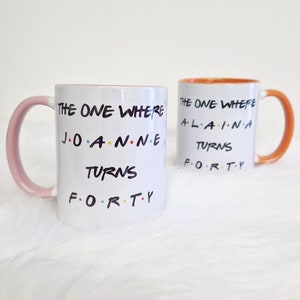 May include: Two white ceramic mugs with the text "THE ONE WHERE... TURNS FORTY" in black, with colorful accents. One mug has a pink handle and rim, the other has an orange handle and rim. The mugs are on a white, fluffy surface.