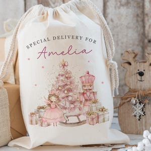 May include: A cream-colored drawstring gift bag with the text "SPECIAL DELIVERY FOR Amelia" in black and pink script. The bag features a pink Christmas tree, fairy, and nutcracker illustration. A wooden reindeer and wrapped gifts are in the background.