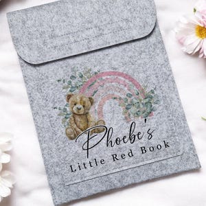 May include: A gray felt book cover with a flap closure. The cover features a watercolor illustration of a teddy bear, a rainbow, and greenery. The text "Phoebe's Little Red Book" is printed below the illustration.