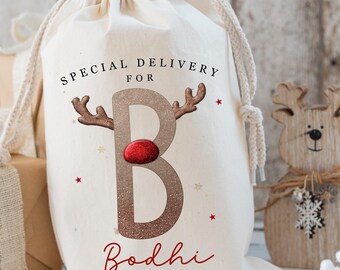 May include: A cream-colored drawstring gift bag with the text "SPECIAL DELIVERY FOR B Bodhi" in red and brown. The letter "B" is designed with reindeer antlers and a red nose. A wooden reindeer decoration is in the background.