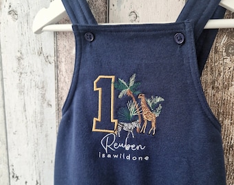 May include: A navy blue baby overall with a large embroidered number one, a giraffe, zebra, and palm trees. The text "Reuben is wild one" is embroidered below the number.