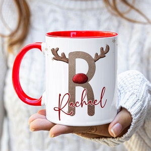 May include: White ceramic mug with a red rim and handle. The mug features a brown reindeer with a red nose and the letter "R" in a glittery gold font. The name "Rachael" is written in red cursive below the reindeer.