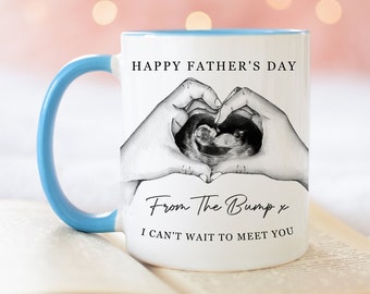 Könnte beinhalten: Eine weiße Keramiktasse mit einem blauen Henkel. Die Tasse zeigt ein Schwarzweißbild eines Ultraschalls mit Händen, die ein Herz darum bilden. Der Text auf der Tasse lautet "Happy Father's Day From The Bump x I can't wait to meet you."