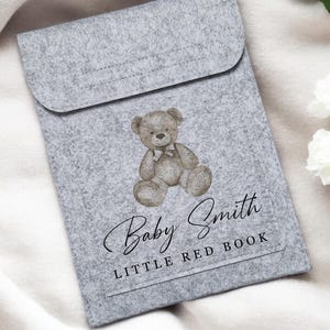 May include: A gray felt book cover with a watercolor illustration of a teddy bear wearing a bow tie. The text "Baby Smith" is written in cursive, with "LITTLE RED BOOK" below. The cover is rectangular with a flap closure.