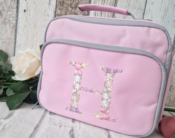 May include: A rectangular light pink lunch bag with a gray zipper trim and a top handle. The front features a large embroidered letter H decorated with a colorful floral pattern consisting of small pink, white, and purple flowers and green leaves.