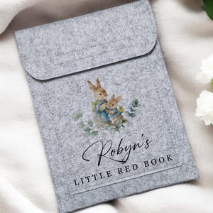 May include: A gray felt book cover with a flap closure. The cover features a watercolor illustration of two rabbits and the text "Robyn's LITTLE RED BOOK" in script and block letters. White flowers are in the background.
