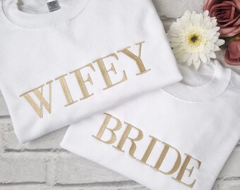 May include: Two white t-shirts with gold embroidery. The top shirt says "WIFEY" and the bottom shirt says "BRIDE".