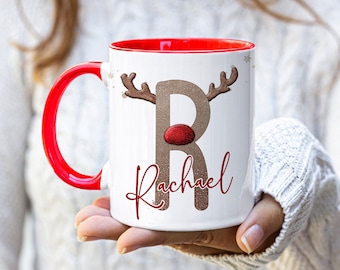 May include: White ceramic mug with a red rim and handle. The mug features a brown reindeer with a red nose and the letter "R" in a glittery gold font. The name "Rachael" is written in red cursive below the reindeer.