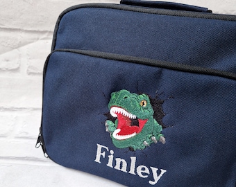 Könnte beinhalten: Eine marineblaue Lunchbox mit schwarzem Reißverschluss und Griff. Die Tasche ist mit einem gestickten grünen Dinosaurier mit roten Zähnen und dem Namen "Finley" in Weiß verziert.