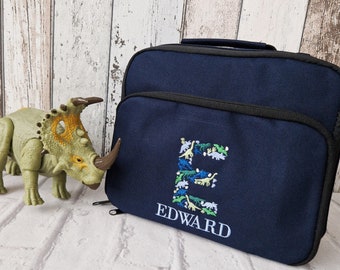 Könnte beinhalten: Eine marineblaue Lunch-Tasche mit Reißverschluss. Die Tasche hat einen schwarzen Griff und ist mit einem dinosaurierförmigen Buchstaben E und dem Namen "EDWARD" bestickt.