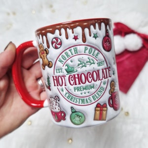 May include: White ceramic mug with a red handle. The mug has a brown chocolate-like glaze dripping down the side. The mug has a festive design with the text "North Pole Est. 1897 Hot Chocolate Premium Christmas Blend".