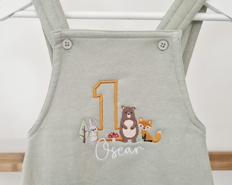 May include: A light green baby overall with a white embroidered number one, a bear, a fox, a rabbit, and trees. The name "Oscar" is embroidered below the number one.