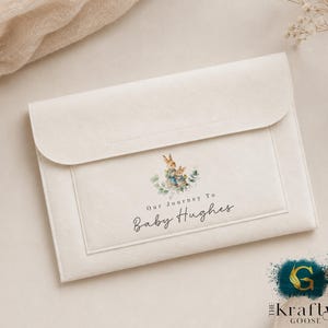 May include: A cream-coloured felt envelope with a flap closure. The front displays a watercolour illustration of two rabbits surrounded by greenery, with the text "Our Journey To Baby Hughes" in script. The Krafty Goose logo is in the bottom right corner.
