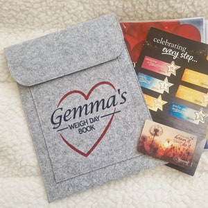 May include: A grey felt pouch with a red heart and the text "Gemma's - Weigh Day Book" printed on it. The pouch is sitting on a white fluffy surface next to a motivational card with the text "You can dream it, do it".