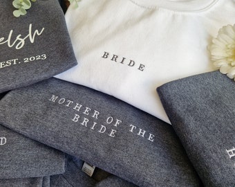 May include: A stack of gray and white t-shirts with embroidered text. The top shirt is white and says "BRIDE". The next shirt is gray and says "MOTHER OF THE BRIDE". The bottom shirt is gray and says "EST. 2023".