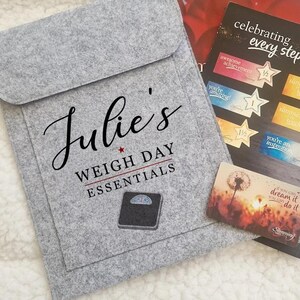 May include: A grey felt pouch with the text "Julie's WEIGH DAY ESSENTIALS" and a scale graphic. Beside it are cards with motivational phrases and star-shaped stickers. The cards read "celebrating every step..."