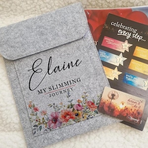 May include: A grey felt pouch with the name "Elaine" and the text "MY SLIMMING JOURNEY" printed on it, along with a floral design. The pouch is next to a card that says "celebrating every step..." and other cards.