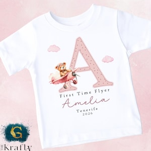 May include: White t-shirt featuring a pink polka-dotted letter "A" and a teddy bear in a pink airplane. The shirt includes the text "First Time Flyer" and the name "Amelia" with "Tenerife 2026" below.