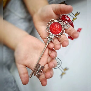 May include: A decorative silver key with a red circular centerpiece. The centerpiece has the number 63 and the text "St Andrews Road". The key is held in a person's hands. The key has the word "HOHOHO" on the key's teeth.
