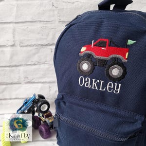 May include: A navy blue backpack featuring a red monster truck embroidery with black wheels and a green flag. The name "Oakley" is embroidered in white below the truck. The bag has a zip closure.