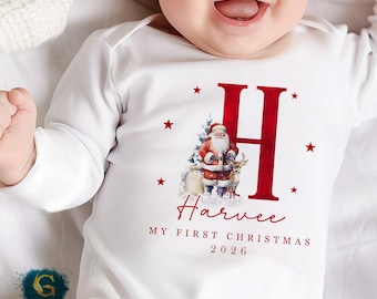 May include: White baby onesie with long sleeves, featuring a red letter "H" and a Christmas-themed graphic of Santa Claus, a reindeer, and a Christmas tree. The text "Harvee, MY FIRST CHRISTMAS 2026" is printed below the graphic.