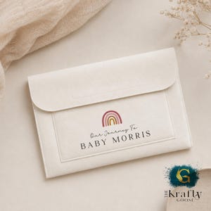 May include: A cream-coloured felt pouch with a flap closure. The front displays a rainbow graphic in pink, yellow, and brown, with the text "Our Journey To BABY MORRIS" printed below. The pouch is a soft, neutral colour.
