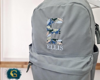 Könnte beinhalten: Grauer Rucksack mit dem gestickten Buchstaben "E", gefüllt mit Dinosaurierformen in verschiedenen Farben. Der Name "ELLIS" ist darunter gestickt. Der Rucksack hat Seitentaschen und schwarze Träger.
