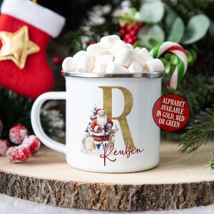 May include: White enamel mug with a gold letter R and the name Reuben. The mug features a Santa Claus illustration with reindeer. The mug is filled with marshmallows and a candy cane.