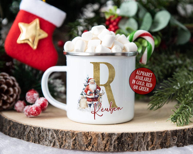 May include: White enamel mug with a gold letter R and the name Reuben. The mug features a Santa Claus illustration with reindeer. The mug is filled with marshmallows and a candy cane.