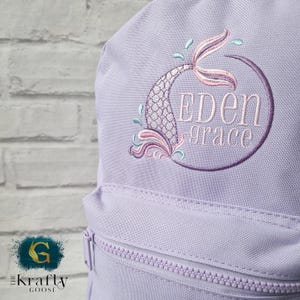 May include: A light purple backpack with a mermaid tail design embroidered in shades of pink, purple, and teal. The name "EDEN grace" is embroidered inside a purple circle. The backpack has a front zipper pocket and a zipper closure.