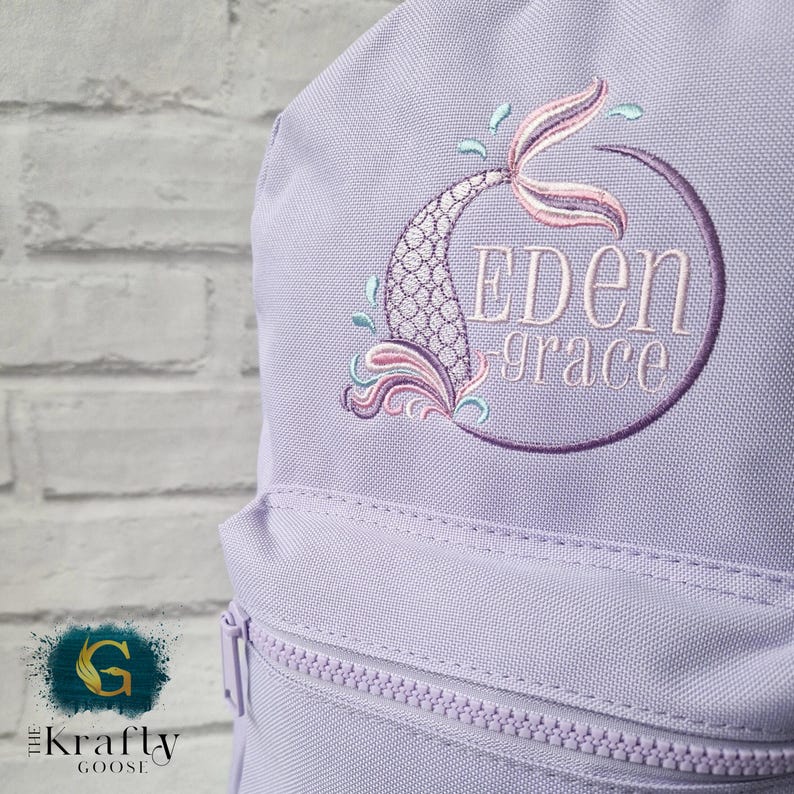 May include: A light purple backpack with a mermaid tail design embroidered in shades of pink, purple, and teal. The name "EDEN grace" is embroidered inside a purple circle. The backpack has a front zipper pocket and a zipper closure.