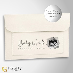 May include: A white felt folder with a pocket on the front. The folder has a label that says "Baby Woods Pregnancy Notes" and a black and white ultrasound image with a watercolor effect. The text "Add Your Own Baby Scan" is written on a white circle.