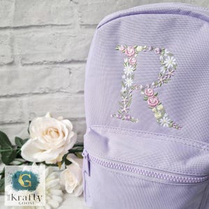 May include: A lavender backpack with a floral embroidered letter "R" on the front. The embroidery features pink, white, and lavender flowers. The backpack has a zippered front pocket and a matching zipper.
