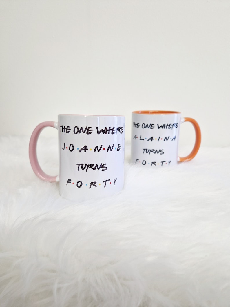 May include: Two white ceramic mugs with the text "THE ONE WHERE... TURNS FORTY" in black, with colorful accents. One mug has a pink handle and rim, the other has an orange handle and rim. The mugs are on a white, fluffy surface.