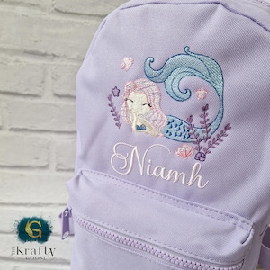 May include: A lilac backpack featuring an embroidered mermaid design. The mermaid has pink hair and a blue tail, with aquatic plants and the name "Niamh" in white script. The backpack has a front zipped pocket.