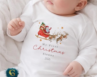 May include: White baby onesie with a Christmas theme. The onesie features a watercolor illustration of Santa Claus in a red sleigh pulled by reindeer, with the text "My First Christmas" and the name "Sienna" and the year "2026".