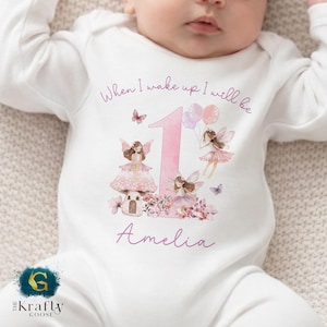 May include: White baby bodysuit featuring a pink number 1, fairies, and the name "Amelia." The text "When I wake up I will be" is above the number. The design includes butterflies, flowers, and balloons.
