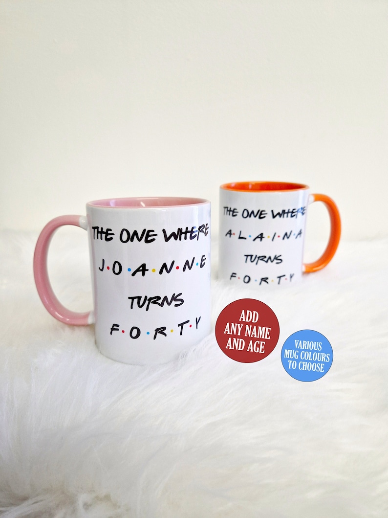May include: Two white ceramic mugs with text, one with a pink handle and interior, the other with an orange handle and interior. Text includes "THE ONE WHERE" and "TURNS FORTY". Additional text on red and blue circles.