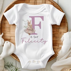 May include: White baby onesie with a pink letter F and a bunny wearing a tutu. The text "F is for Felicity" is printed below the letter.