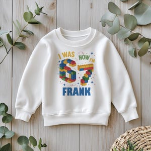 May include: White sweatshirt with the text "I WAS 6, NOW I'M 7 FRANK" in colorful block letters. The number 6 and 7 are designed to look like building blocks. The sweatshirt is a crew neck style.