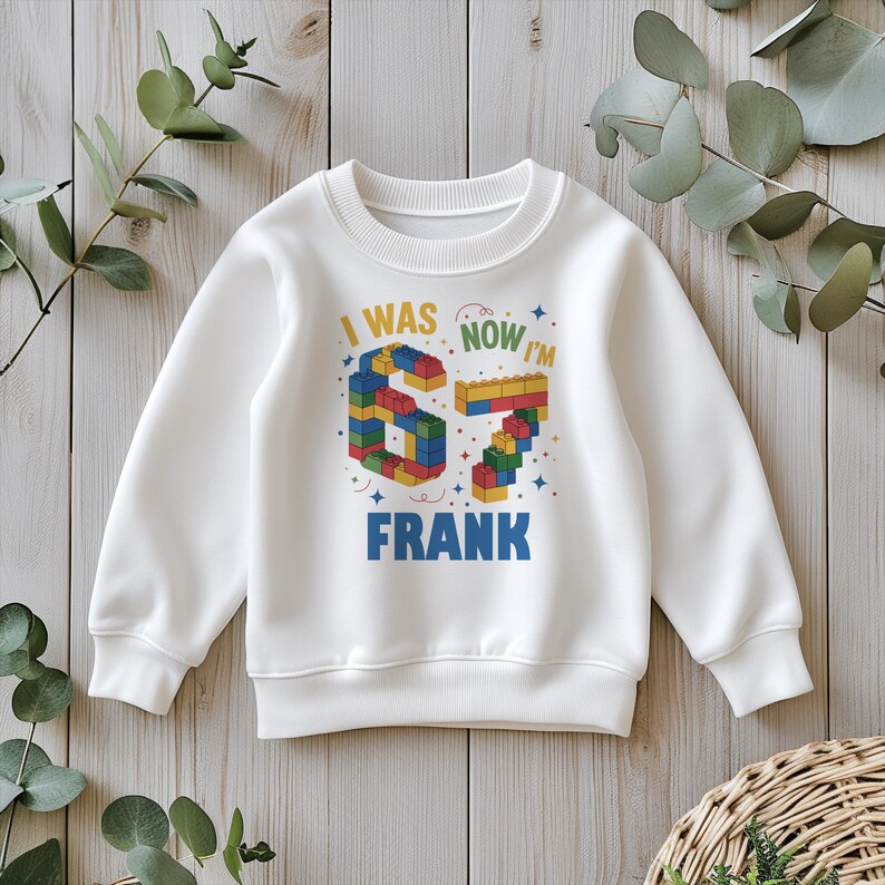 May include: White sweatshirt with the text "I WAS 6, NOW I'M 7 FRANK" in colorful block letters. The number 6 and 7 are designed to look like building blocks. The sweatshirt is a crew neck style.