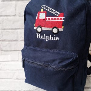 May include: A navy blue backpack with a red fire engine embroidered on the front. The backpack has a zipped pocket on the front and the name "Ralphie" embroidered below the fire engine.