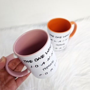 May include: Two white ceramic mugs with black text and colorful accents. One mug has a pink interior and handle, the other has an orange interior and handle. The text on the mugs reads "THE ONE WHERE JOANNA TURNS FORTY."