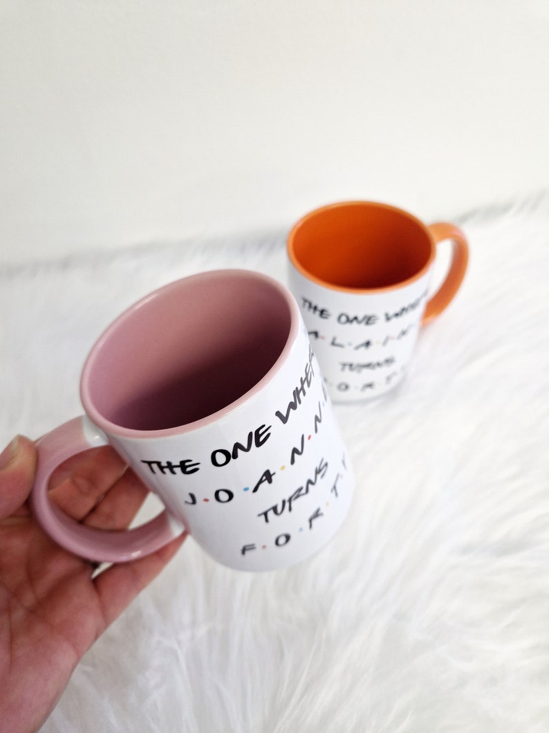 May include: Two white ceramic mugs with black text and colorful accents. One mug has a pink interior and handle, the other has an orange interior and handle. The text on the mugs reads "THE ONE WHERE JOANNA TURNS FORTY."