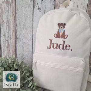 May include: Cream-colored backpack with a front pocket and a top handle. The backpack features an embroidered brown teddy bear with a blue bow tie and the name "Jude" in brown thread. The backpack has a zipper closure.
