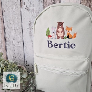 May include: A pale green backpack with a front zipped pocket. It features an embroidered design of a bear, fox, and rabbit, with the name "Bertie" in navy blue. The backpack has a top handle and adjustable straps.