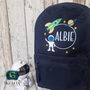 May include: Navy blue backpack with a front zippered pocket. The backpack features an embroidered design of a green rocket, a planet with rings, stars, and an astronaut inside a white circle. The name "ALBIE" is embroidered in white.