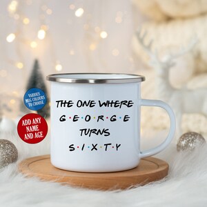 May include: White enamel mug with a silver rim, featuring the text "THE ONE WHERE GEORGE TURNS SIXTY" in black, with the name in colorful letters. The mug sits on a wooden coaster, with decorative elements in the background.