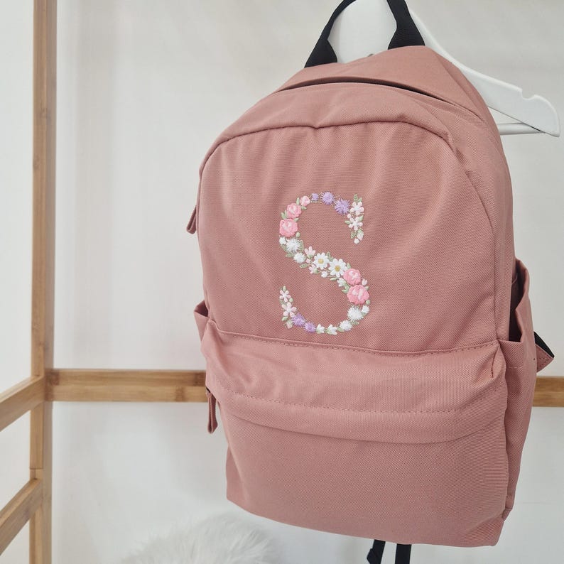 Puede incluir: Una mochila rosa empolvado con una "S" bordada floral en la parte delantera. El bordado presenta flores rosas, blancas y moradas. La mochila tiene un bolsillo delantero, bolsillos laterales y correas negras.